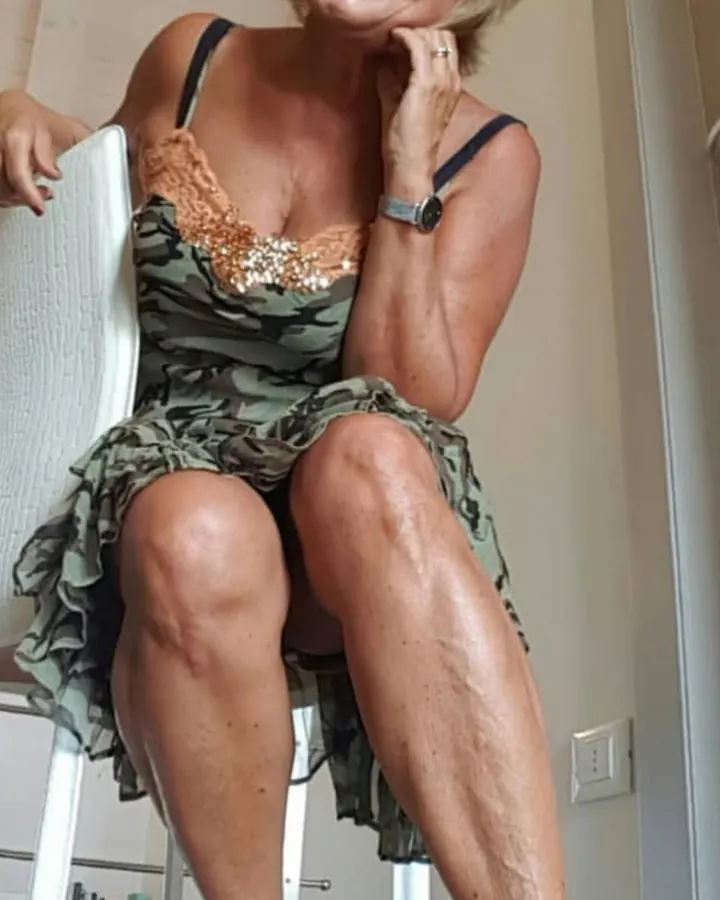 Femme aux cheveux blonds courts, en robe noire à sequins, assise sur un canapé beige, souriant à la caméra.