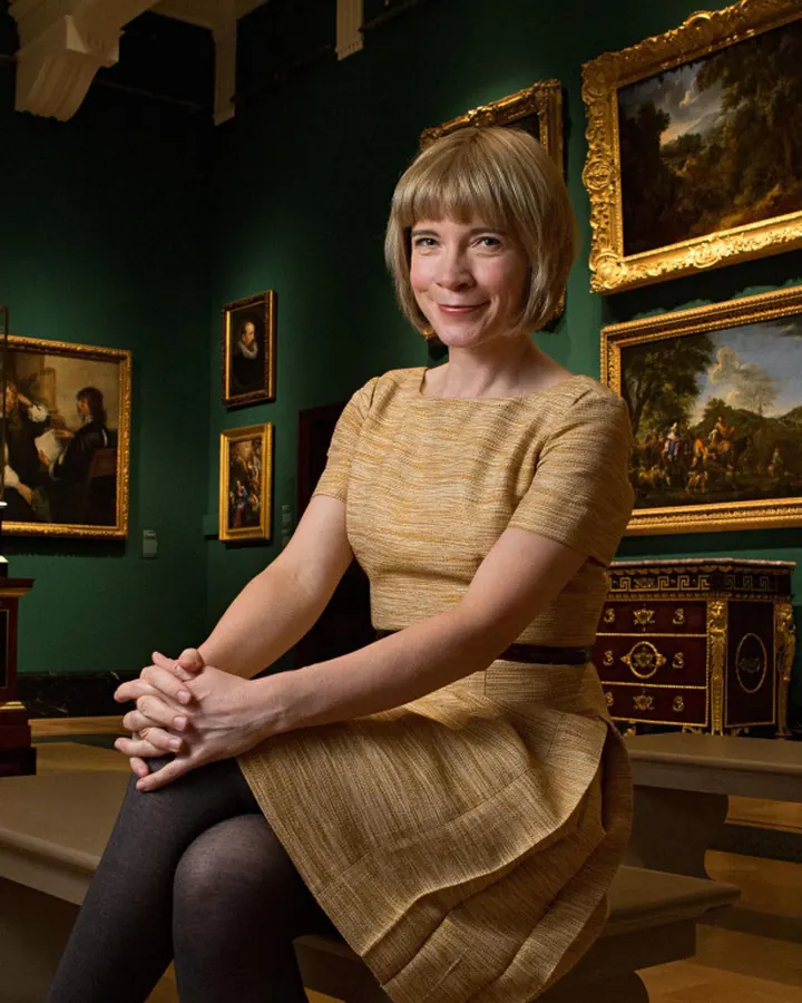 Femme aux cheveux blonds courts, en robe beige, assise sur un banc dans une salle de musée, entourée de tableaux dorés.