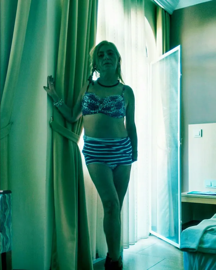 Femme aux cheveux blonds, vêtue d'un haut à motifs léopard et d'un short rayé bleu et blanc, debout près d'une fenêtre, tenant une serviette, dans une pièce aux tons verts.