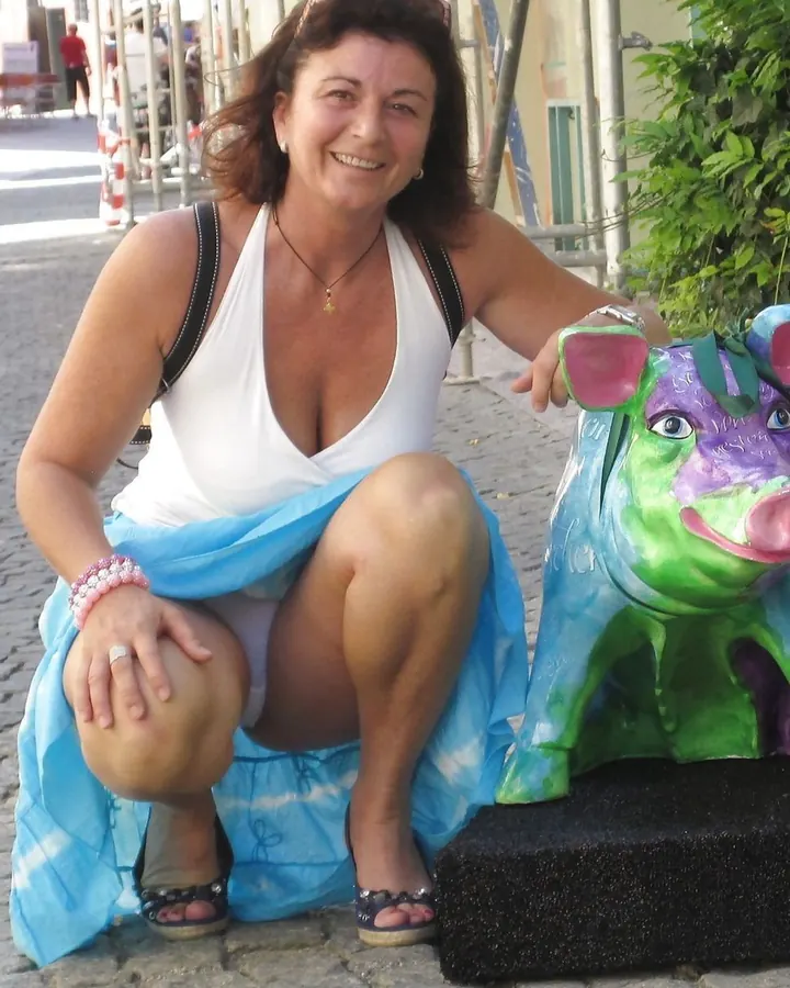 Femme châtain aux yeux bleus souriante, assise sur un trottoir pavé, vêtue d'un haut blanc profondément décolleté et d'une jupe bleue, près d'une sculpture colorée de cochon.