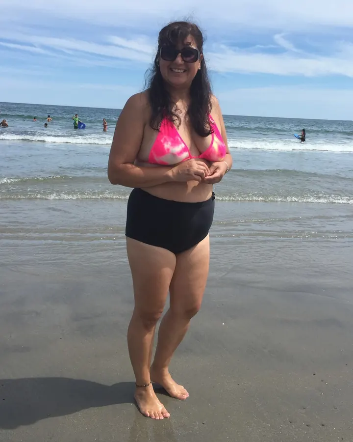 Femme aux cheveux bruns-noirs, souriante, assise sur une plage, vêtue d'un haut de maillot de bain rose et blanc et d'un bas noir, les bras croisés sur son ventre, les pieds nus dans le sable.