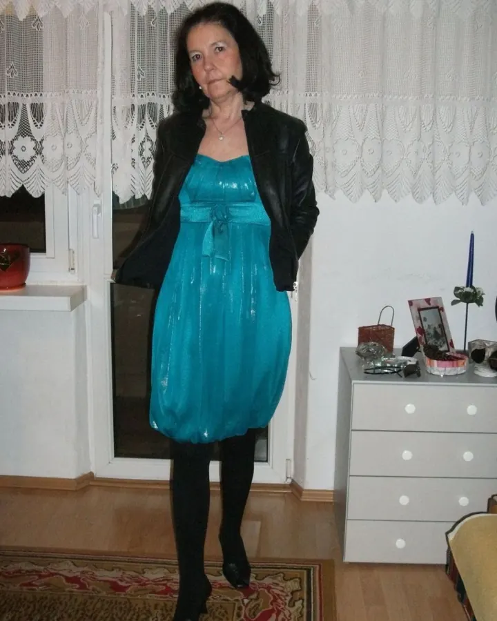 Femme aux cheveux bruns-noirs, vêtue d'une robe turquoise brillante, d'une veste en cuir noir, collants noirs, chaussures noires, debout dans une pièce avec rideaux blancs et meubles clairs.