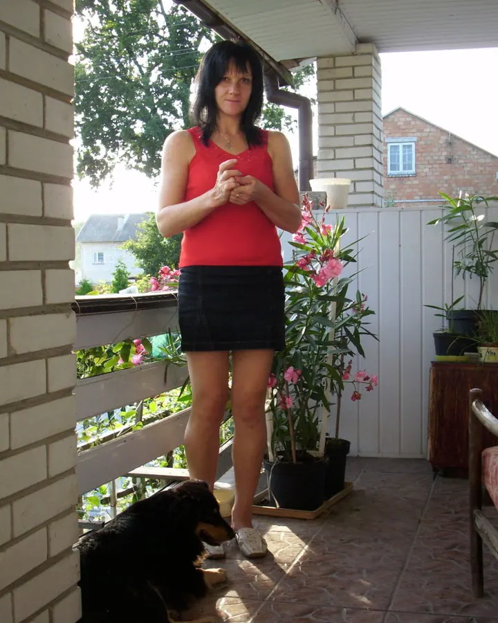 Femme aux cheveux noirs portant un haut rouge et une jupe noire, debout sur une terrasse avec un chien noir à ses pieds.