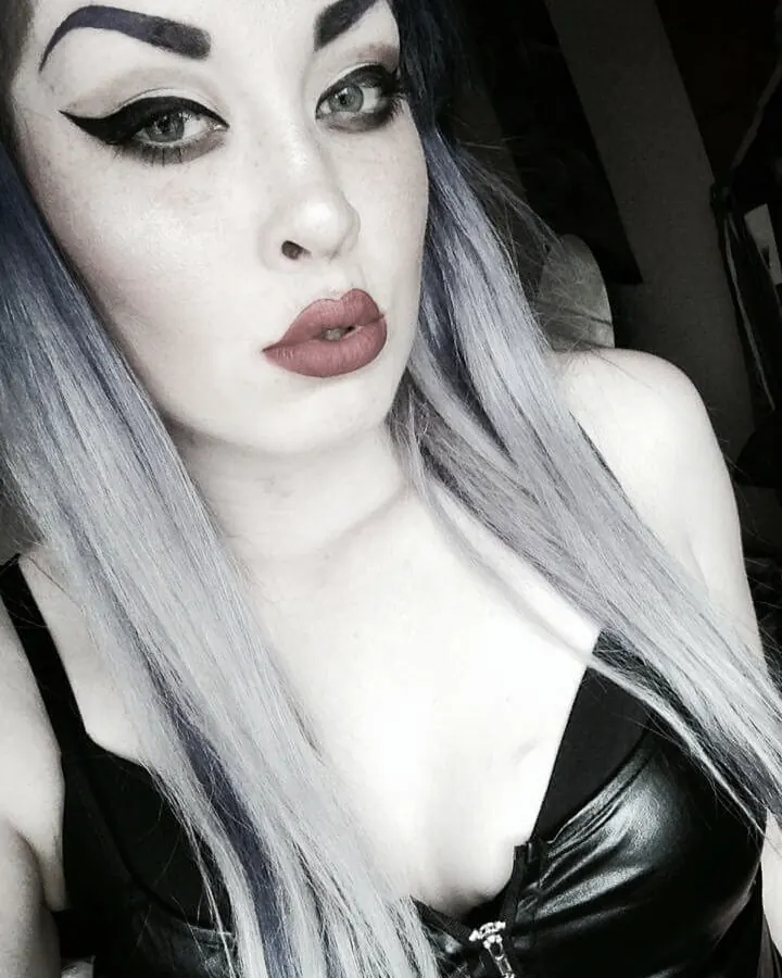 Femme aux cheveux gris et maquillage prononcé, portant un haut noir en cuir, regardant l'appareil photo avec intensité.