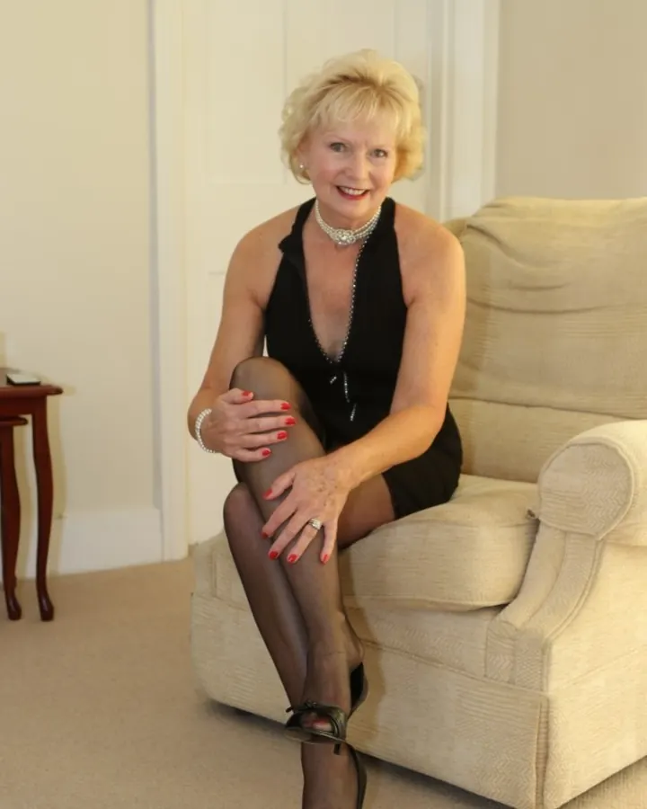 Femme aux cheveux blonds platine, portant un haut noir sans manches et une jupe noire, assise sur un canapé noir avec des collants noirs et des bottines noires à talons.