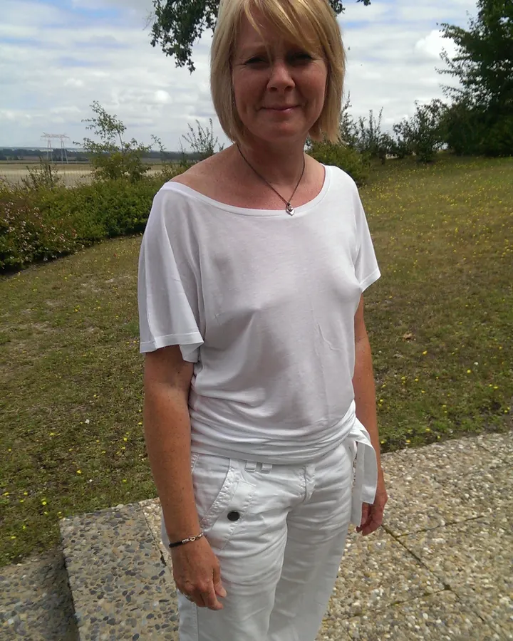 Femme aux cheveux blonds courts, portant un t-shirt blanc ample et un pantalon blanc, debout sur un chemin pavé dans un champ verdoyant sous un ciel nuageux.