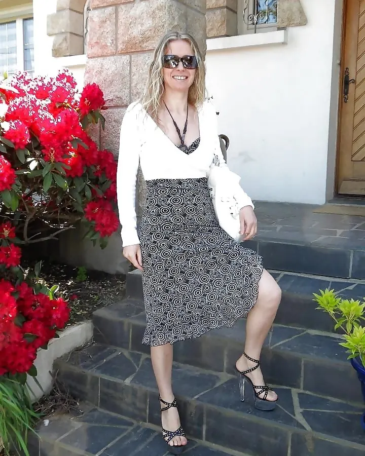 Femme aux cheveux blonds platine, portant un haut noir sans manches et une jupe noire, assise sur un canapé noir avec des collants noirs et des bottines noires à talons.