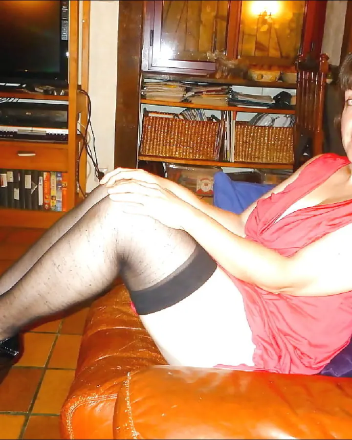 Femme aux cheveux châtain clair en robe noire, collant noir, chaussures à talons, assise sur un canapé orange, main sur la cuisse, regard direct