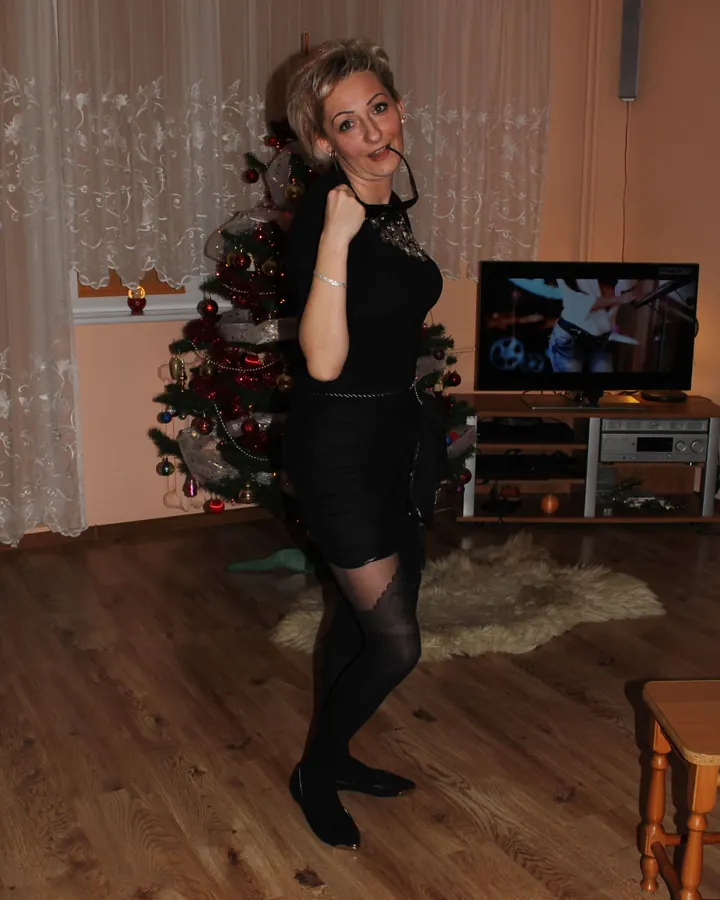 Femme blonde en robe noire, collant noir, chaussures à talons, debout près d'un sapin décoré, lunettes de soleil sur la tête, sourire pétillant