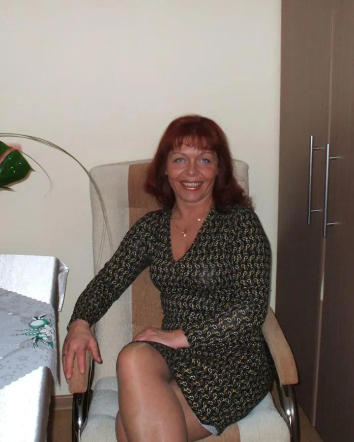 Femme aux cheveux roux souriante, assise dans un fauteuil beige, vêtue d'une robe noire, collant résille, chaussures à talons, regard vers le côté