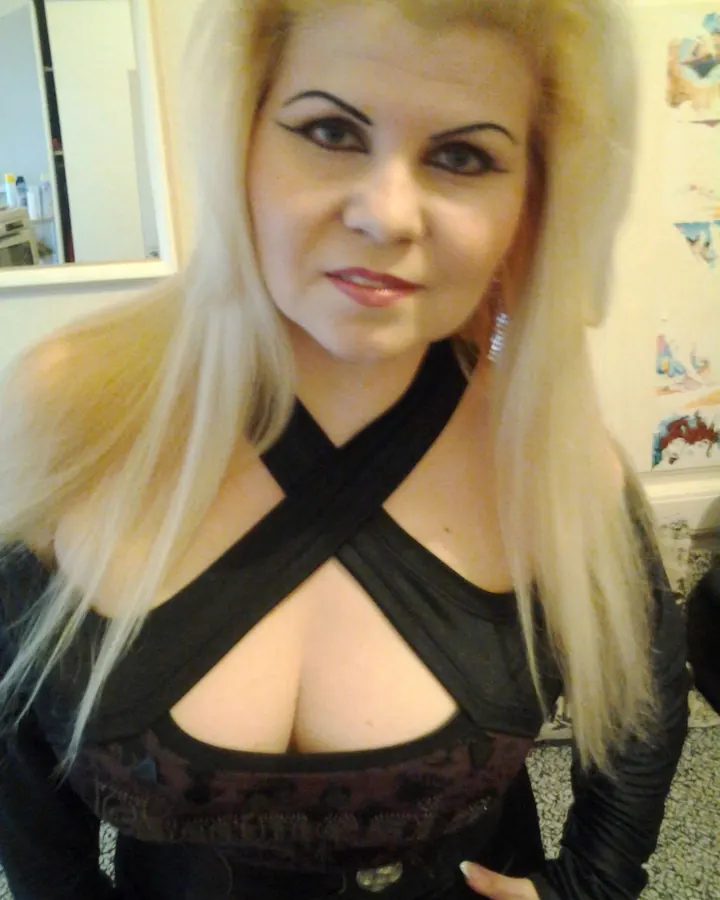 Femme aux cheveux blonds platine en tenue noire, maquillage prononcé, regard intense, pose frontale