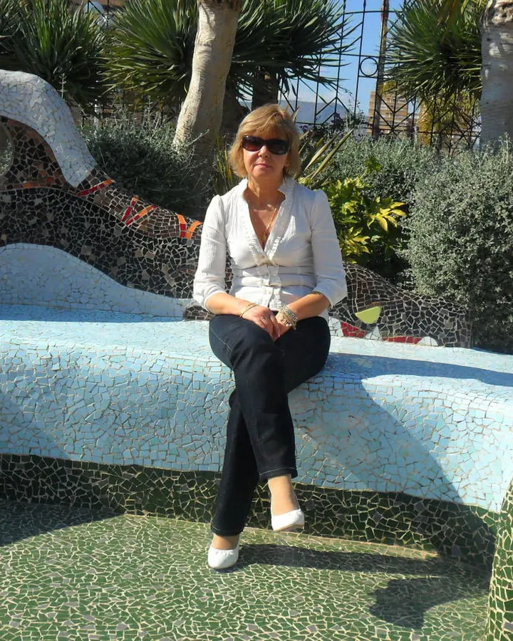 Femme blonde assise sur un banc en mosaïque colorée, vêtue d'une chemise blanche et de jeans, lunettes de soleil, sourire discret