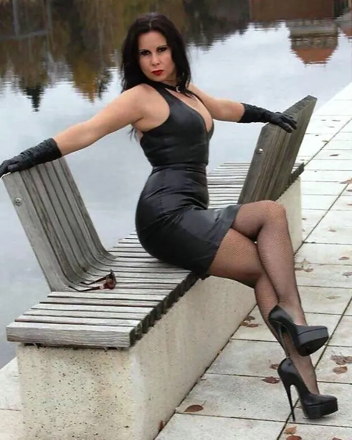 Femme aux cheveux bruns-noirs assise sur un banc en bois près d’un lac, vêtue d’une robe noire ajustée, de gants noirs et de talons hauts.