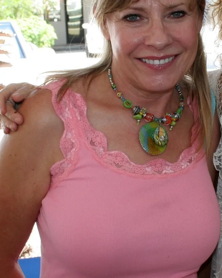 Femme aux cheveux châtains portant un haut rose à dentelle et un collier coloré, souriante en extérieur.