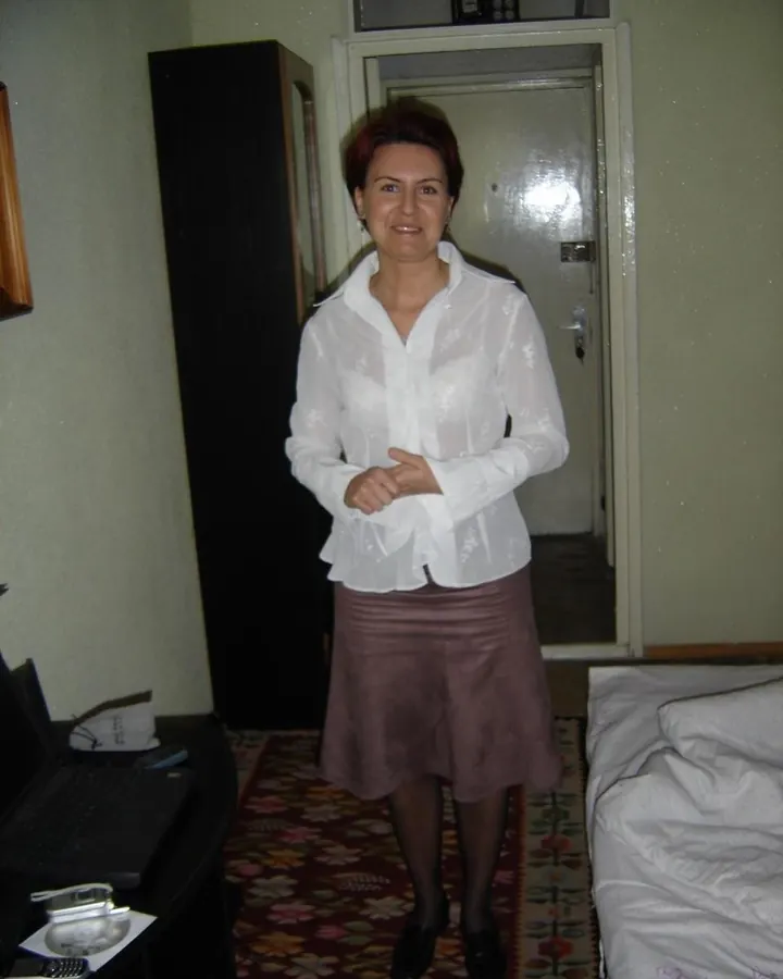 Femme aux cheveux roux courts, portant une chemise blanche transparente et une jupe marron, souriant à la caméra dans une chambre.