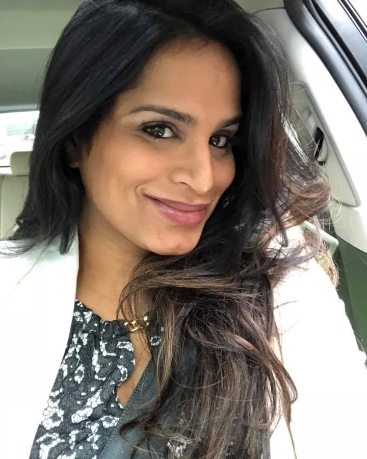Femme aux cheveux noirs, souriante, portant une veste blanche et une blouse à motifs, assise dans une voiture, avec des reflets sur le pare-brise.