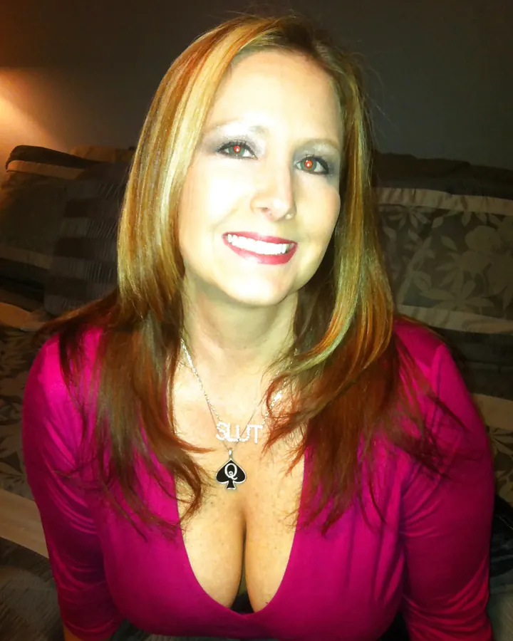 Femme aux cheveux blonds, souriante, portant un haut rose vif décolleté, assise sur un canapé sombre avec un collier orné du mot 'SLUT' et un pendentif trèfle.