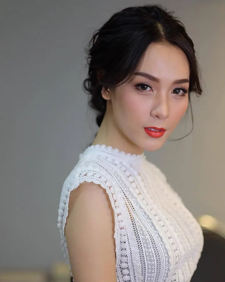 Portrait d'une femme asiatique aux cheveux noirs coiffés en chignon, portant un haut blanc en dentelle, avec un maquillage soigné et un regard direct, sur fond gris.