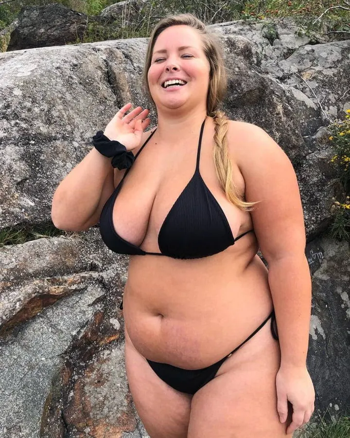 Femme souriante en maillot de bain noir triangle, debout sur des rochers, tenant sa main près de son visage, avec une queue de cheval blonde.