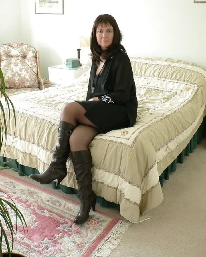 Femme aux cheveux bruns portant une robe noire et des bottes hautes, assise sur un lit avec couvre-lit beige.