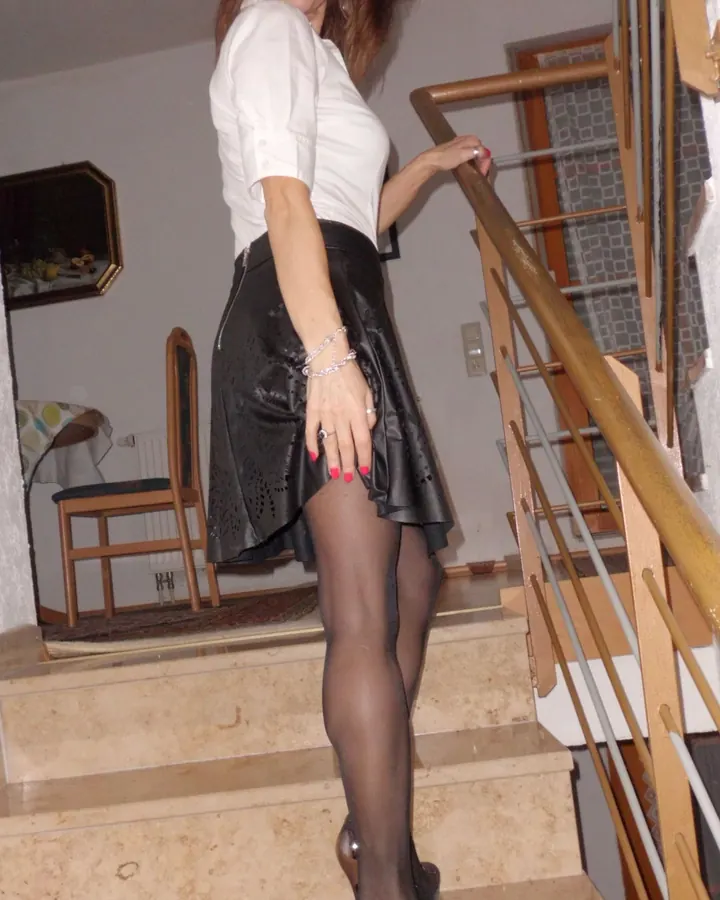 Femme aux cheveux châtains portant un haut noir et un short noir, avec des collants opaques, posant sur un escalier en marbre.