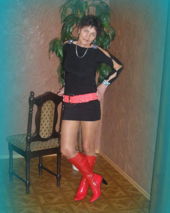 Femme aux cheveux courts noirs en haut noir à épaules dénudées, jupe courte noire, ceinture rose, bottes rouges, posant près d'une chaise ancienne.