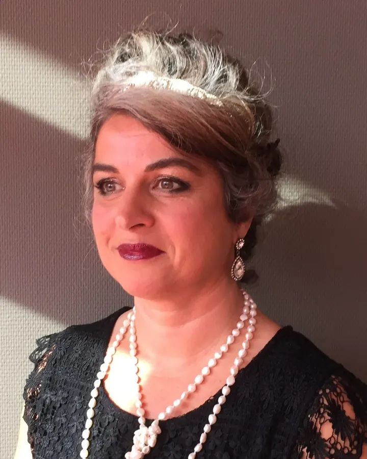 Femme aux cheveux gris, coiffure élégante, portant une tiare et un collier de perles, regardant de côté sous un éclairage contrasté.