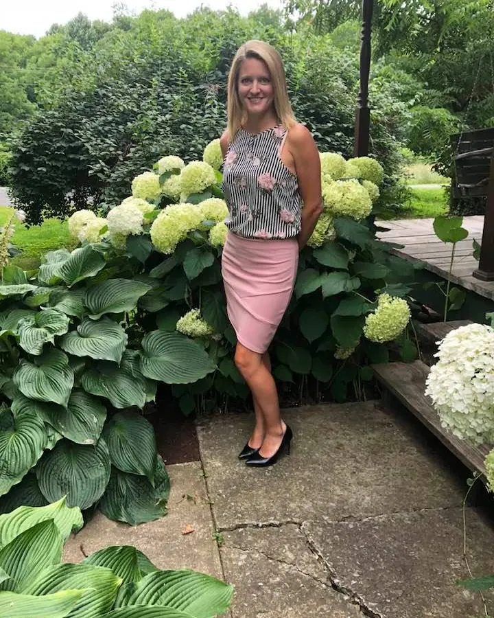 Femme aux cheveux blonds, portant un haut noir et une jupe rose, debout devant un massif de fleurs blanches et vertes, souriant à la caméra.