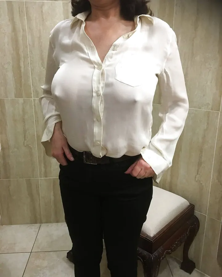 Femme aux cheveux noirs, portant un corset noir, des gants noirs, des bottes en cuir noir, assise sur un canapé noir, tenant une cravache, dans une pièce avec un sol rouge.