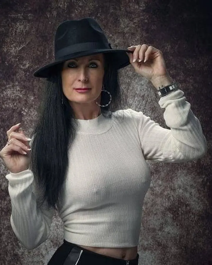 Femme aux cheveux longs noirs, portant un chapeau noir et un haut blanc, tenant le bord de son chapeau avec une main, l’autre levée, devant un fond sombre texturé.