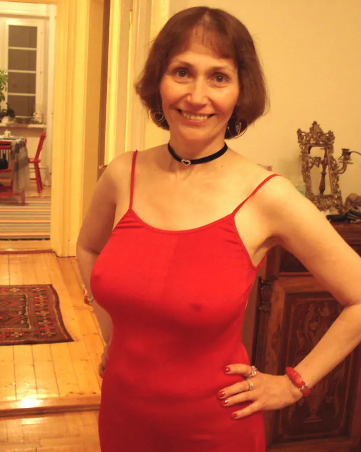 Femme aux cheveux bruns portant une robe rouge, debout dans un couloir, main sur la hanche, souriant.