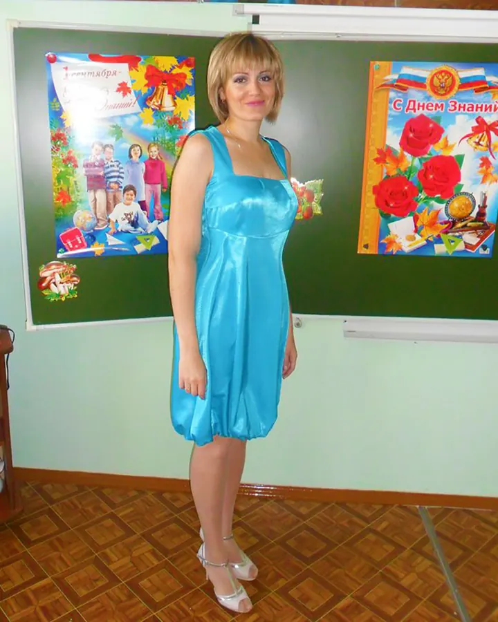 Femme aux cheveux blonds en robe noire, assise sur un canapé bleu, les jambes croisées, regardant l'appareil.