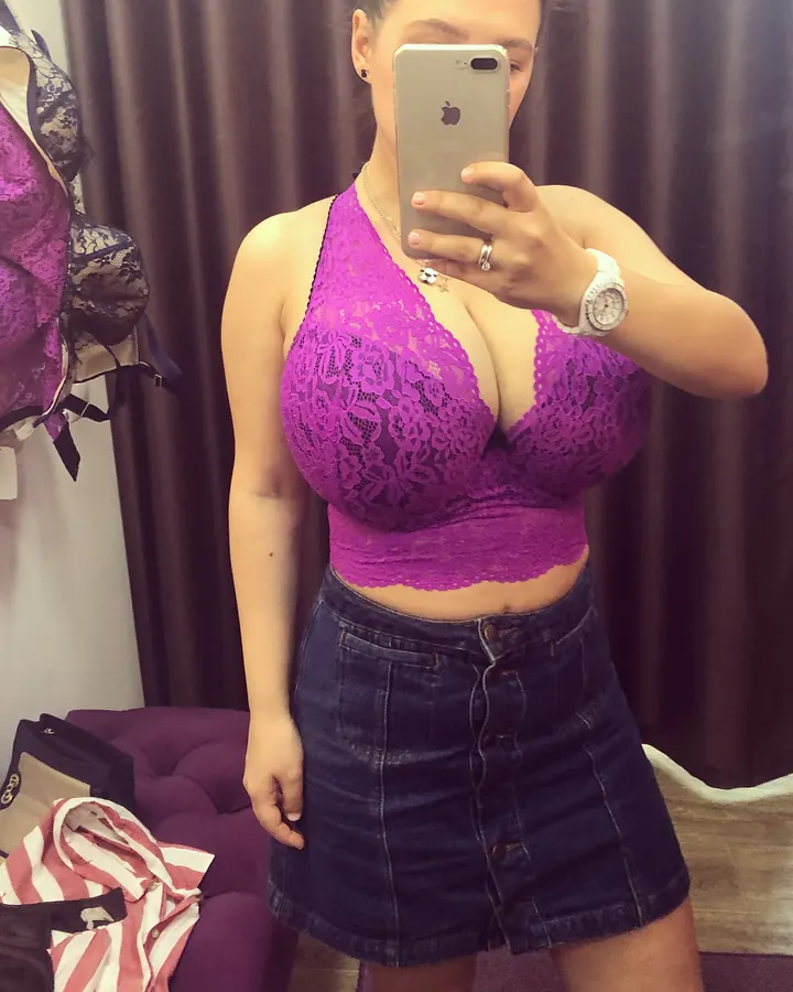Femme aux cheveux bruns foncés portant un haut en dentelle violette et une jupe en jean, prenant un selfie devant un miroir dans une chambre.