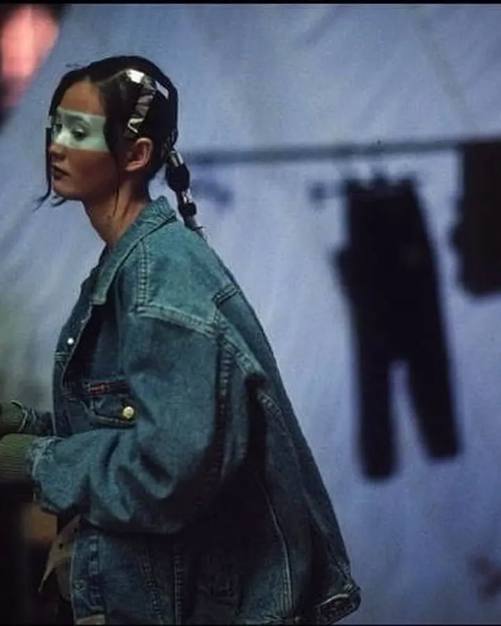 Femme aux cheveux noirs portant une veste en jean et un masque blanc sur les yeux, dans un environnement sombre avec des vêtements suspendus en arrière-plan.