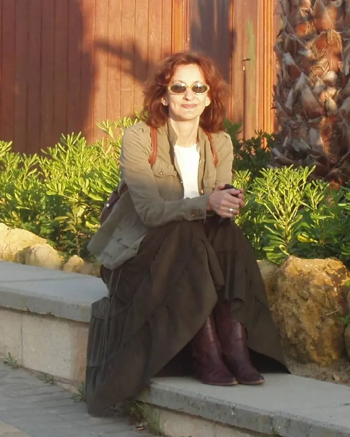 Femme aux cheveux roux, vêtue d'une veste kaki et d'une jupe noire, assise sur un mur de pierre dans un jardin verdoyant.