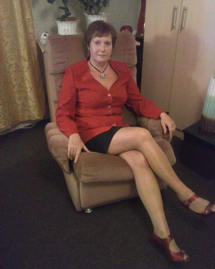 Femme aux cheveux bruns courts, vêtue d'une chemise rouge, assise dans un fauteuil beige dans un salon intime.