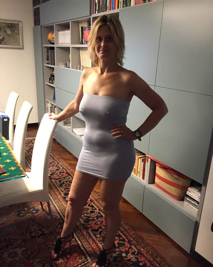 Femme aux cheveux blonds, vêtue d'une robe grise moulante, posant dans une pièce avec une bibliothèque bleue.