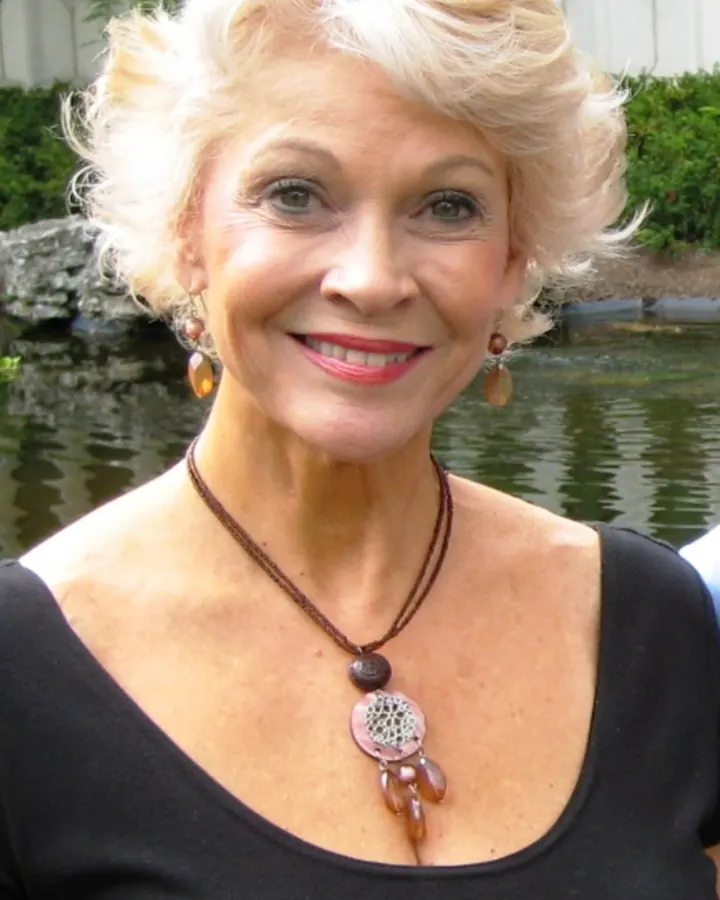 Femme blonde aux cheveux courts, souriante, portant un collier ethnique, devant un arrière-plan végétal.