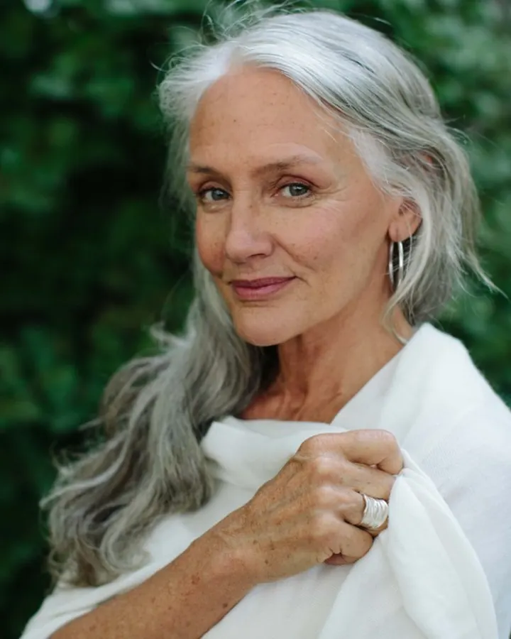 Femme aux cheveux gris argentés, vêtue d'un tissu blanc, souriant légèrement devant un arrière-plan de feuillage vert.