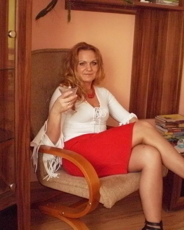 Femme assise dans un fauteuil, en haut blanc et jupe rouge, tenant un verre, dans un salon.
