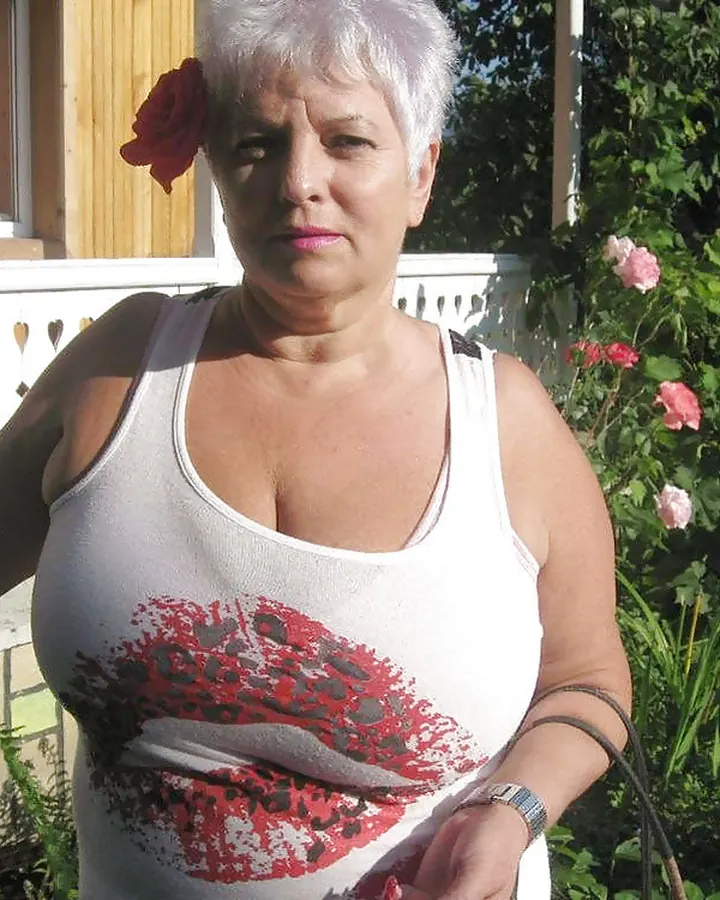 Femme en haut blanc, avec une fleur rouge dans les cheveux gris courts, debout dans un jardin fleuri.