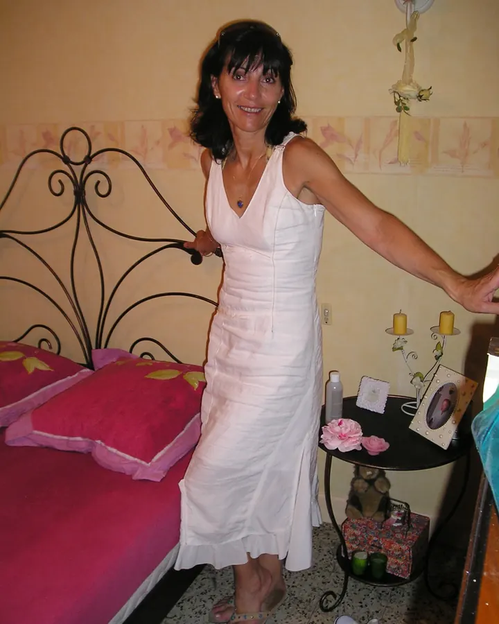Femme en robe blanche, debout près d'un lit à tête de lit métallique, souriant vers l'appareil photo.